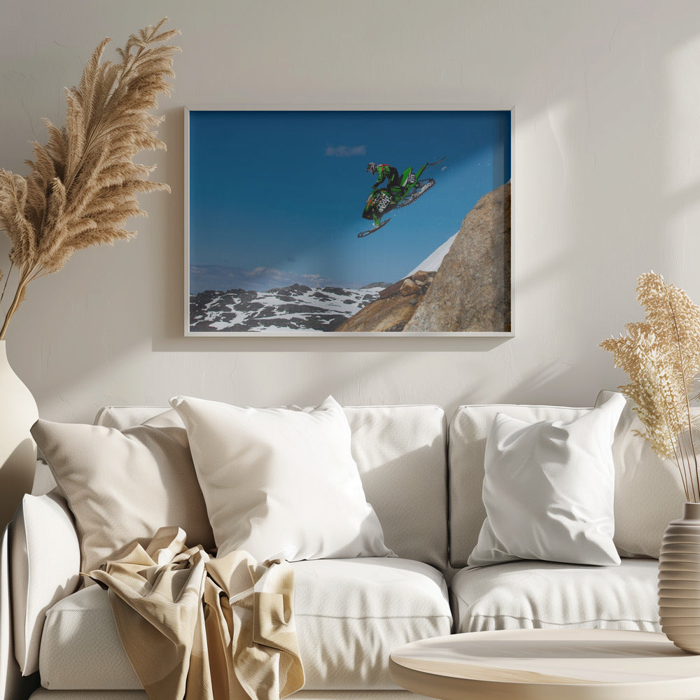 A dynamic action shot of a person in full gear launching a green snowmobile into the air off a rocky, snow-covered cliff, set against a backdrop of a vivid blue sky and distant snowy mountains. The image is enclosed in a silver frame. Wall Art