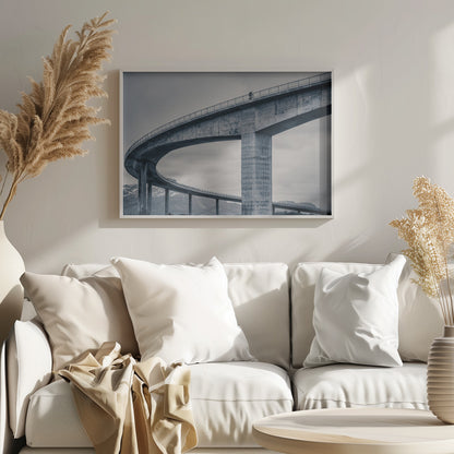 A black and white photograph of a lone cyclist riding across the top level of a massive, curving concrete bridge, which sweeps across the frame. In the background, a lower level of the bridge and distant mountains are visible under a cloudy sky. Decor