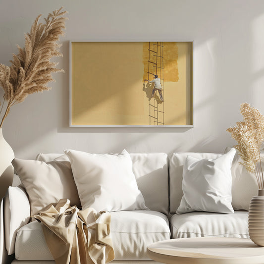 A man viewed from behind stands on a rope ladder, painting a large, bright yellow wall a slightly darker shade of yellow, casting a stark shadow in the bright sun. The image is presented within a silver frame. Decor