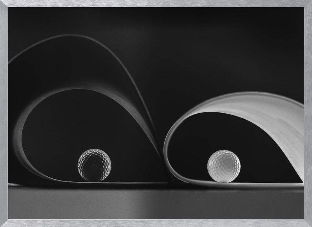A framed, black and white abstract photograph featuring two golf balls nestled within the graceful curves of a single sheet of paper, creating a striking interplay of light, shadow, and form against a dark background. Decor