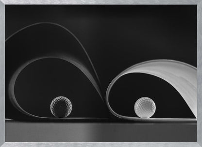 A framed, black and white abstract photograph featuring two golf balls nestled within the graceful curves of a single sheet of paper, creating a striking interplay of light, shadow, and form against a dark background. Decor