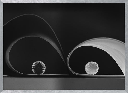 A framed, black and white abstract photograph featuring two golf balls nestled within the graceful curves of a single sheet of paper, creating a striking interplay of light, shadow, and form against a dark background. Decor