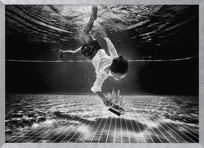 A surreal black and white underwater photograph of a child in a white shirt and shorts diving towards the bottom of a tiled pool to retrieve a miniature model of a sailing ship. Artwork