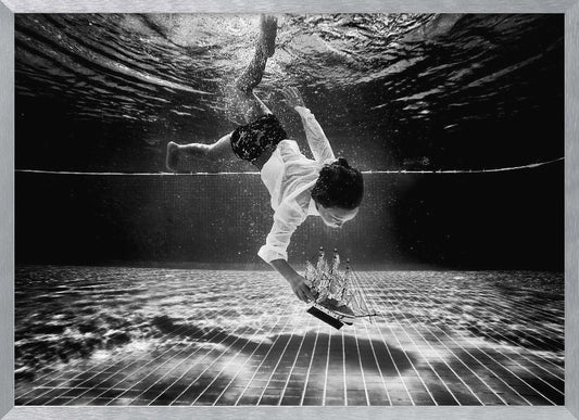 A surreal black and white underwater photograph of a child in a white shirt and shorts diving towards the bottom of a tiled pool to retrieve a miniature model of a sailing ship. Artwork