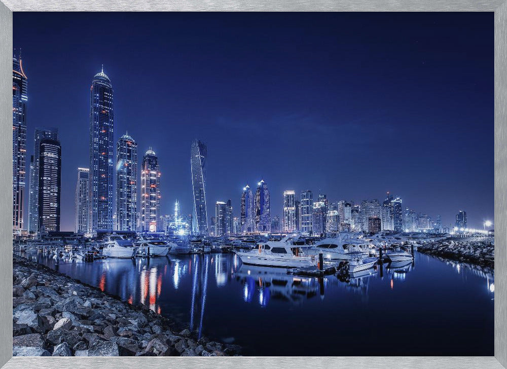 A stunning nighttime photograph of the Dubai Marina skyline, with illuminated skyscrapers towering against a deep blue sky. In the foreground, luxury yachts are moored in the calm harbor, their white hulls and the city's lights creating vibrant reflections on the water's surface. The shot is taken from a rocky embankment. Decor