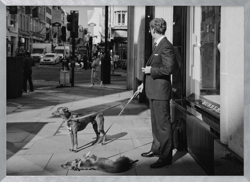 A black and white street photograph of a well-dressed man in a suit standing on a city sidewalk outside a storefront. He holds the leashes for two dogs; one is standing alert, and the other is lying on its back on the pavement. The background is a busy city street with traffic and pedestrians. Print