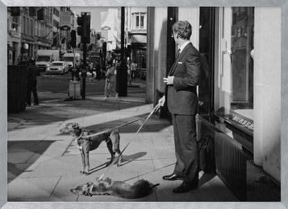 A black and white street photograph of a well-dressed man in a suit standing on a city sidewalk outside a storefront. He holds the leashes for two dogs; one is standing alert, and the other is lying on its back on the pavement. The background is a busy city street with traffic and pedestrians. Print