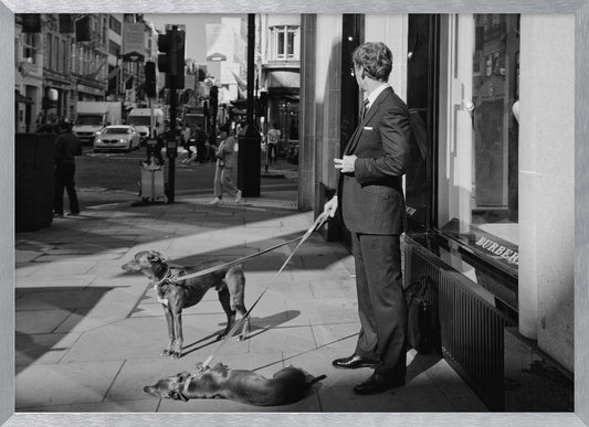 A black and white street photograph of a well-dressed man in a suit standing on a city sidewalk outside a storefront. He holds the leashes for two dogs; one is standing alert, and the other is lying on its back on the pavement. The background is a busy city street with traffic and pedestrians. Print