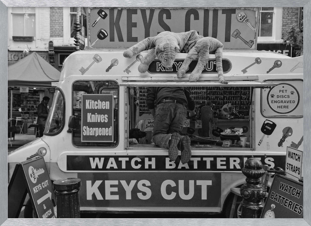 A quirky black and white photograph of a mobile key-cutting van on a city street. A man in jeans is seen from behind, kneeling inside the service window. On the roof, a large stuffed tiger lies stretched out, peering down. The van is covered in signs advertising 'KEYS CUT', 'WATCH BATTERIES', and 'Kitchen Knives Sharpened'. Print