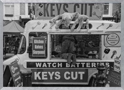 A quirky black and white photograph of a mobile key-cutting van on a city street. A man in jeans is seen from behind, kneeling inside the service window. On the roof, a large stuffed tiger lies stretched out, peering down. The van is covered in signs advertising 'KEYS CUT', 'WATCH BATTERIES', and 'Kitchen Knives Sharpened'. Print
