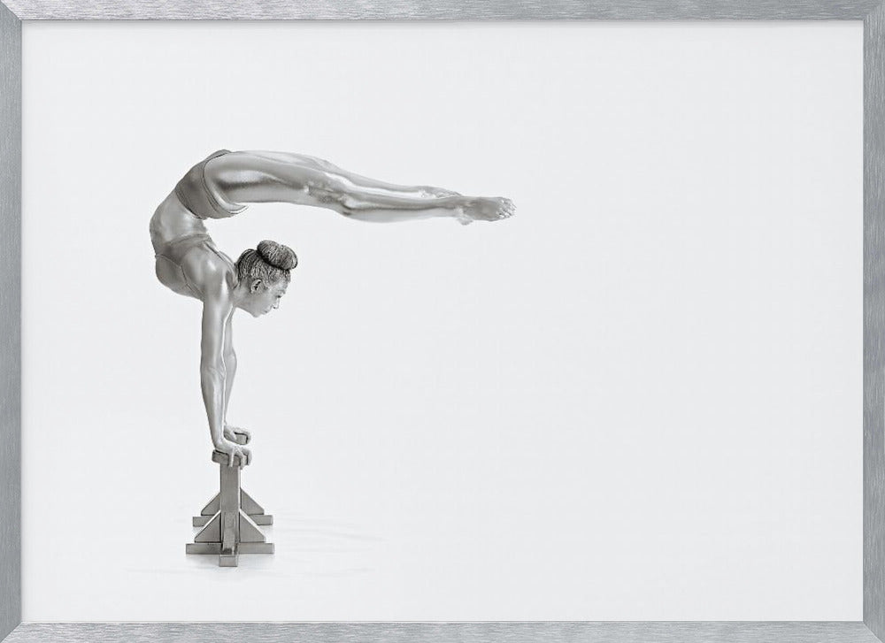 A framed black and white photograph of a female gymnast with a metallic silver-painted body, balancing in a difficult handstand pose on small blocks against a stark white background. Her back is arched and her legs are extended straight out behind her, parallel to the ground. Artwork