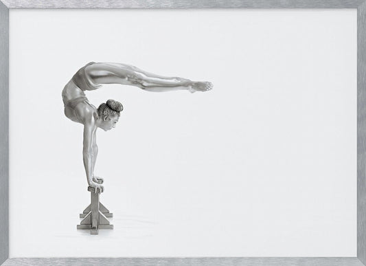 A framed black and white photograph of a female gymnast with a metallic silver-painted body, balancing in a difficult handstand pose on small blocks against a stark white background. Her back is arched and her legs are extended straight out behind her, parallel to the ground. Artwork