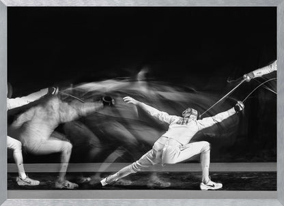 A framed, dramatic black and white photograph using long exposure to capture a fencer in motion. The fencer, in full white gear, lunges forward, their form crisp at the end of the movement while a ghostly blur trails behind, illustrating their rapid advance against a black background. Decor