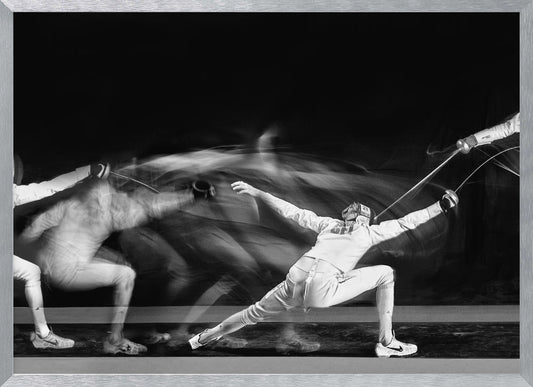 A framed, dramatic black and white photograph using long exposure to capture a fencer in motion. The fencer, in full white gear, lunges forward, their form crisp at the end of the movement while a ghostly blur trails behind, illustrating their rapid advance against a black background. Decor
