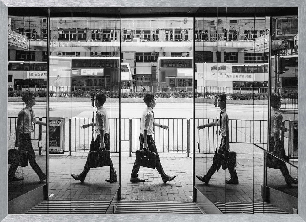 A black and white street photograph showing a man in a shirt and tie carrying a briefcase, his walking motion captured in a time-lapse effect across multiple reflective glass panels. The reflections create a sequence of his stride, set against the backdrop of a busy city street with buses and buildings also reflected in the glass. Decor