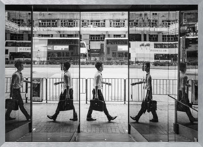 A black and white street photograph showing a man in a shirt and tie carrying a briefcase, his walking motion captured in a time-lapse effect across multiple reflective glass panels. The reflections create a sequence of his stride, set against the backdrop of a busy city street with buses and buildings also reflected in the glass. Decor