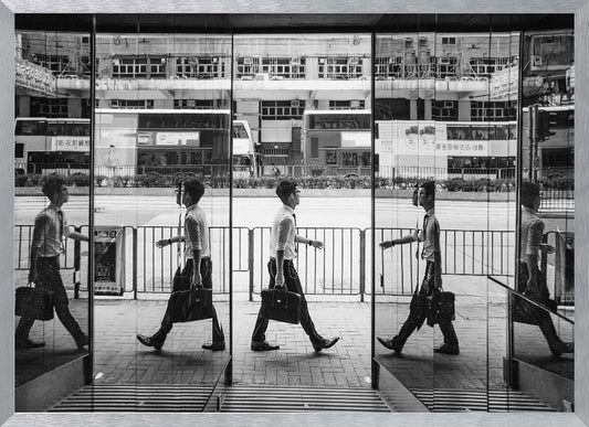 A black and white street photograph showing a man in a shirt and tie carrying a briefcase, his walking motion captured in a time-lapse effect across multiple reflective glass panels. The reflections create a sequence of his stride, set against the backdrop of a busy city street with buses and buildings also reflected in the glass. Decor