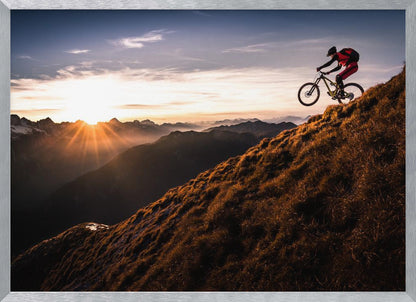 A mountain biker in mid-air, jumping down a steep grassy mountain ridge at sunrise. The sun is low on the horizon, casting a golden glow and long sunbeams over a vast, layered mountain range in the background. Poster
