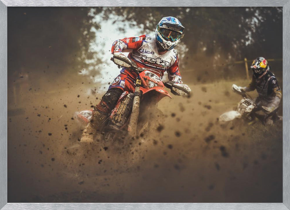 An action shot of a motocross rider in red, white, and blue gear, speeding through a dusty turn on a dirt track, kicking up a large cloud of mud and debris. Another racer is visible in the background. The image is presented within a silver frame. Print