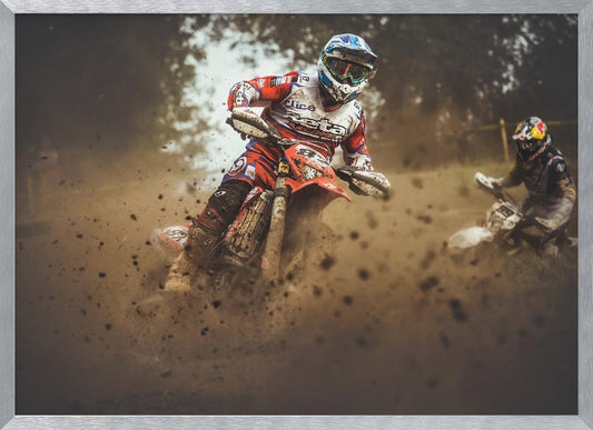 An action shot of a motocross rider in red, white, and blue gear, speeding through a dusty turn on a dirt track, kicking up a large cloud of mud and debris. Another racer is visible in the background. The image is presented within a silver frame. Print