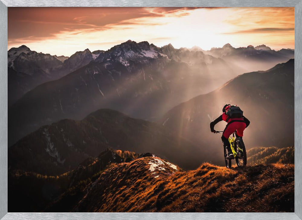 A mountain biker in red and black gear pauses on a grassy, sunlit ridgeline, looking out at a vast mountain range with snow-capped peaks as the sun sets behind them, casting dramatic rays of light through the valleys. Poster