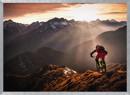 A mountain biker in red and black gear pauses on a grassy, sunlit ridgeline, looking out at a vast mountain range with snow-capped peaks as the sun sets behind them, casting dramatic rays of light through the valleys. Poster