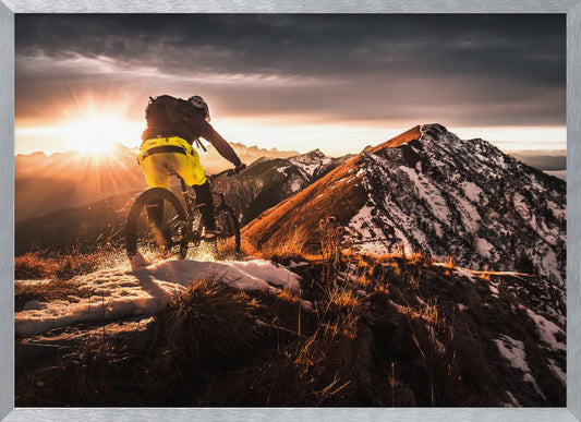 A mountain biker seen from behind rides along a narrow, snowy trail on a mountain ridge during a dramatic sunset, with the sun flaring brightly and a vast, snow-capped mountain range in the background. Decor