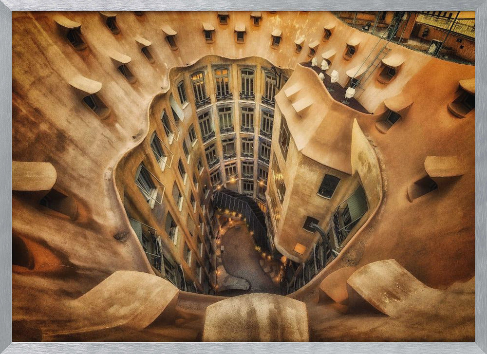 A high-angle, downward view of the inner courtyard of an architecturally unique building, likely Casa Milà by Gaudí. The warm, sandy-colored walls curve and undulate organically, surrounding the multi-story structure with its many windows. The courtyard below is lit, casting a warm glow. Decor