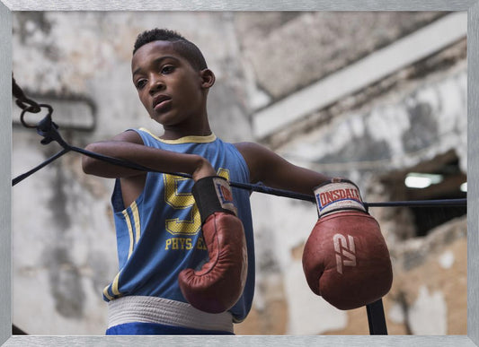 A young Black boy in a blue jersey and red boxing gloves leans pensively against the ropes of a boxing ring, looking away with a thoughtful expression against a blurred, weathered background. Wall Art