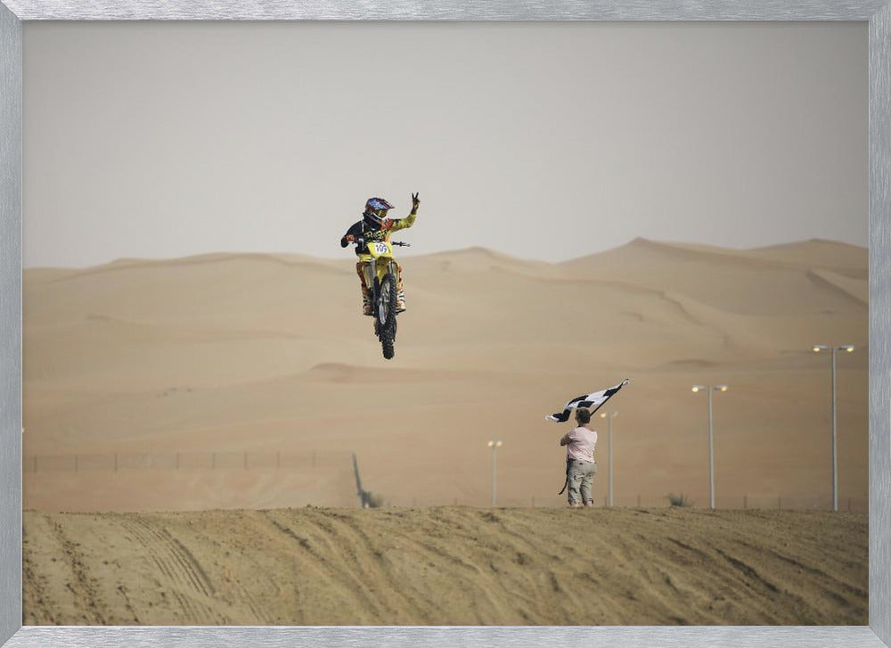 A motocross rider on a yellow dirt bike is captured mid-air during a jump in a desert landscape, flashing a peace sign with their left hand. In the background, rolling sand dunes stretch under a hazy sky, and to the right, a person on a hill waves a black and white checkered flag. Print