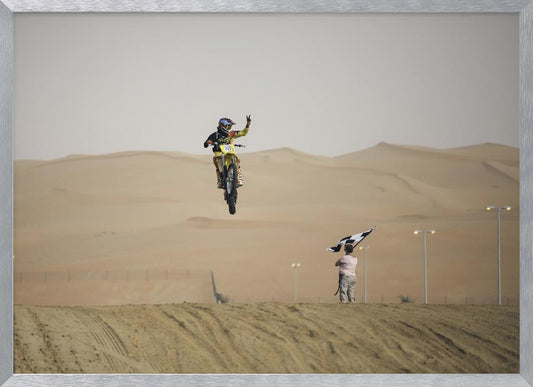 A motocross rider on a yellow dirt bike is captured mid-air during a jump in a desert landscape, flashing a peace sign with their left hand. In the background, rolling sand dunes stretch under a hazy sky, and to the right, a person on a hill waves a black and white checkered flag. Print