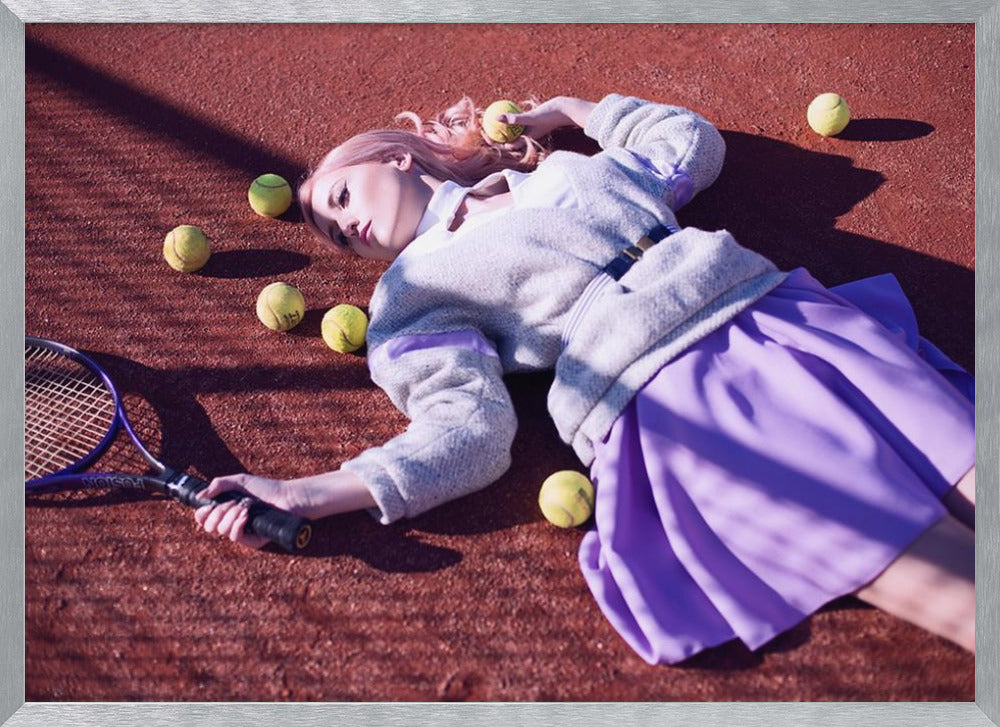 A high-angle shot of a young woman with pink hair lying on a red clay tennis court, surrounded by yellow tennis balls. She wears a grey sweater, a white collared shirt, and a purple pleated skirt, and holds a tennis racket in one hand. The scene is framed with a silver border. Wall Art