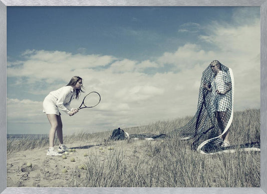 A surreal photograph of two women on a grassy sand dune. One woman is dressed in white tennis attire, bent over and ready to hit a ball with her racket. The other woman stands opposite her, entangled and draped in a large tennis net. The scene is under a cloudy blue sky and has a desaturated, cool-toned color palette, framed in silver. Decor