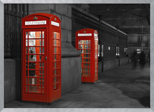 A selective color photograph of a city street at night, framed in silver. Two iconic red telephone booths are the only objects in color, standing out against the black and white background of the cobblestone sidewalk and iron fence. Artwork