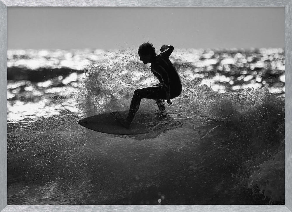 A black and white action shot of a surfer silhouetted against the sparkling ocean. The surfer is crouched low on their board, carving through a wave and creating a dramatic spray of water. The photo is enclosed in a silver frame. Print