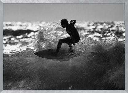 A black and white action shot of a surfer silhouetted against the sparkling ocean. The surfer is crouched low on their board, carving through a wave and creating a dramatic spray of water. The photo is enclosed in a silver frame. Print