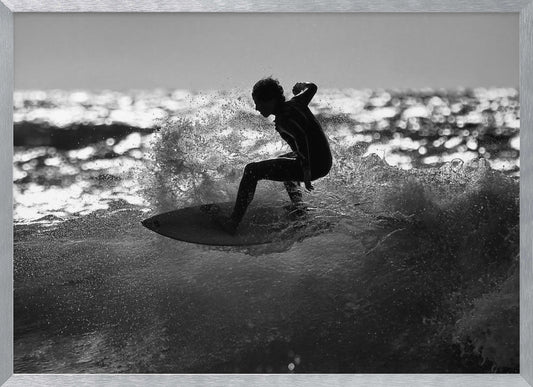 A black and white action shot of a surfer silhouetted against the sparkling ocean. The surfer is crouched low on their board, carving through a wave and creating a dramatic spray of water. The photo is enclosed in a silver frame. Print