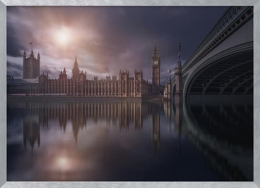 A dramatic, atmospheric photograph of the Houses of Parliament and Big Ben in London, reflected in the still waters of the River Thames. A bright sun breaks through dark, moody clouds, casting a warm light on the scene, with a stone bridge visible to the right. The image is enclosed in a silver frame. Print