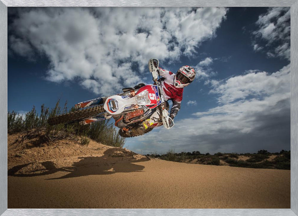 A low-angle action shot of a motocross rider on a dirt bike catching major air over a sand dune against a cloudy blue sky. The image is presented within a silver frame. Decor