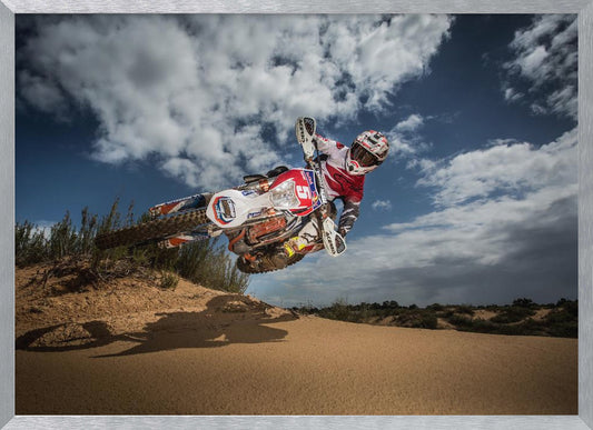 A low-angle action shot of a motocross rider on a dirt bike catching major air over a sand dune against a cloudy blue sky. The image is presented within a silver frame. Decor