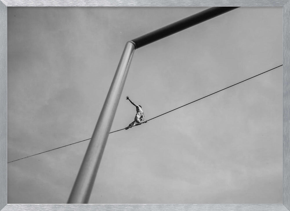A black and white, low-angle photograph of a person balancing while sitting on a tightrope high in the sky. A large metal pole is in the foreground, creating a strong diagonal line. The image is framed in silver. Decor