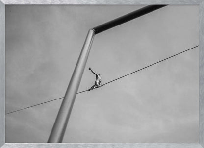 A black and white, low-angle photograph of a person balancing while sitting on a tightrope high in the sky. A large metal pole is in the foreground, creating a strong diagonal line. The image is framed in silver. Decor