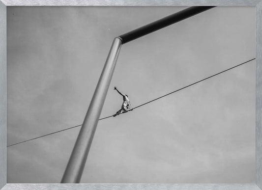 A black and white, low-angle photograph of a person balancing while sitting on a tightrope high in the sky. A large metal pole is in the foreground, creating a strong diagonal line. The image is framed in silver. Decor
