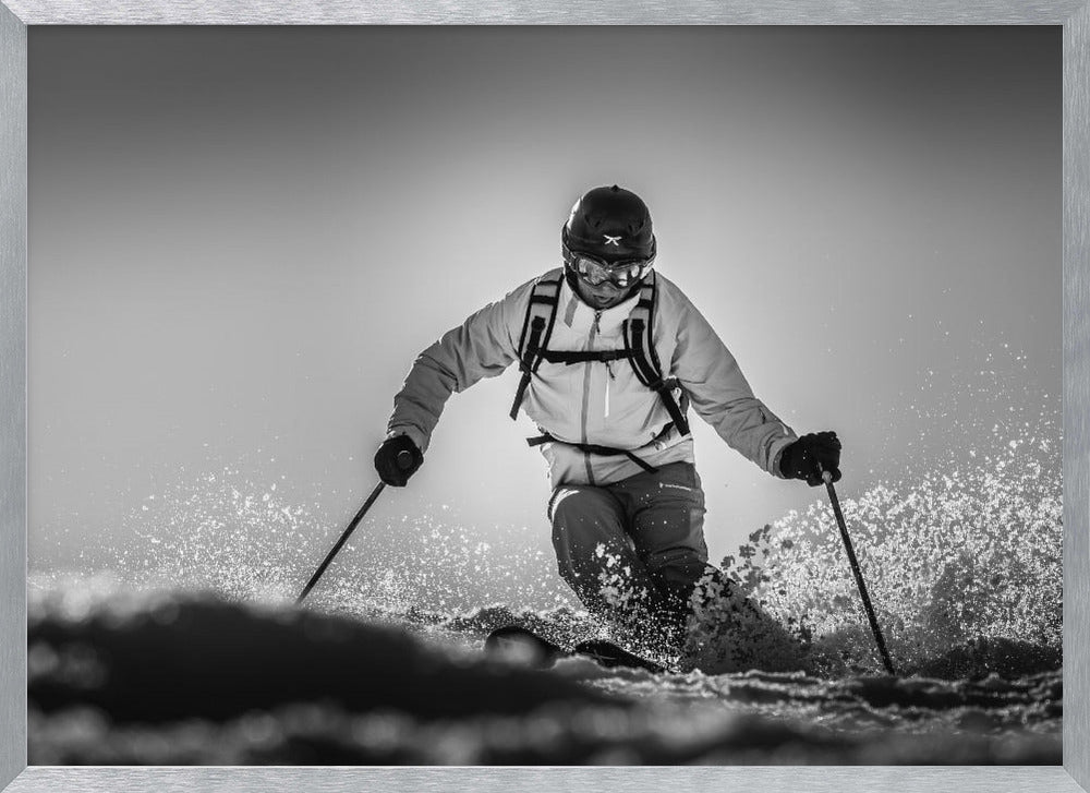 A black and white action photograph of a skier in full gear, leaning into a turn and spraying powder snow. The skier is backlit by the sun, creating a high-contrast, dramatic effect. Print