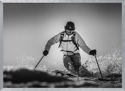 A black and white action photograph of a skier in full gear, leaning into a turn and spraying powder snow. The skier is backlit by the sun, creating a high-contrast, dramatic effect. Print