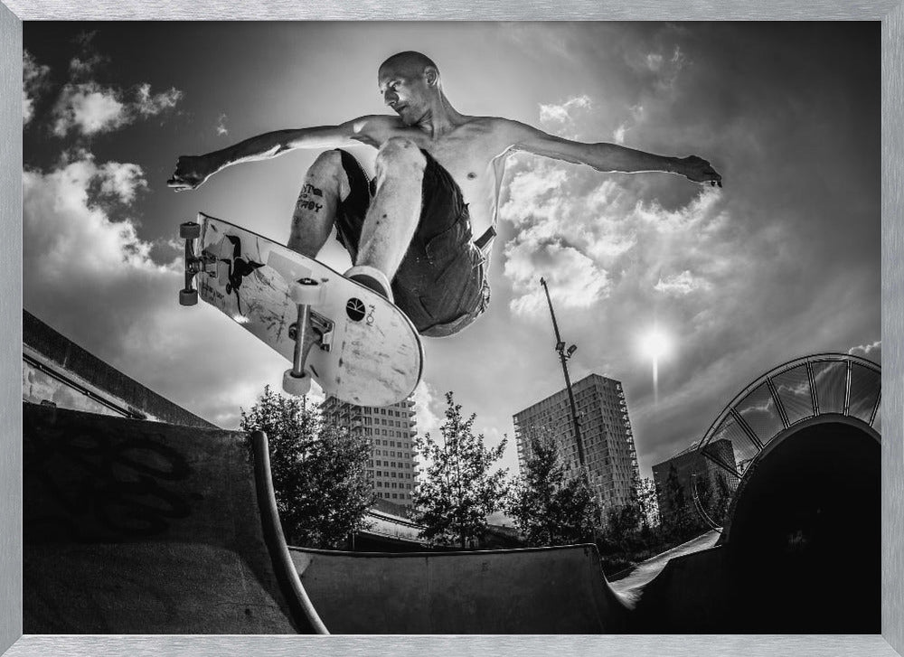 A dramatic low-angle, black and white photograph of a shirtless skateboarder caught in mid-air above a skatepark ramp. His arms are spread wide for balance as the sun flares brightly behind him, set against a cloudy urban sky with high-rise buildings in the distance. The image is enclosed in a silver frame. Wall Art