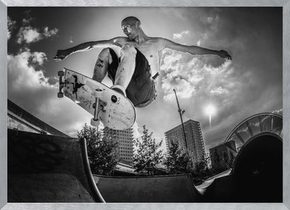 A dramatic low-angle, black and white photograph of a shirtless skateboarder caught in mid-air above a skatepark ramp. His arms are spread wide for balance as the sun flares brightly behind him, set against a cloudy urban sky with high-rise buildings in the distance. The image is enclosed in a silver frame. Wall Art