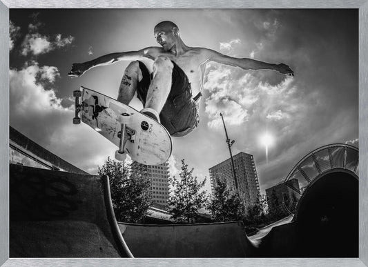 A dramatic low-angle, black and white photograph of a shirtless skateboarder caught in mid-air above a skatepark ramp. His arms are spread wide for balance as the sun flares brightly behind him, set against a cloudy urban sky with high-rise buildings in the distance. The image is enclosed in a silver frame. Wall Art
