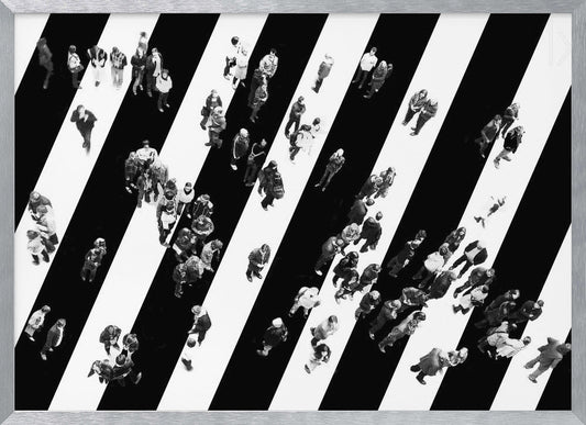 A striking black and white aerial photograph of dozens of people walking across a large, diagonally striped crosswalk. The high-contrast, graphic pattern of black and white stripes dominates the scene, with the small figures of pedestrians scattered across it, creating a sense of movement and urban anonymity. The image is presented within a simple silver frame. Print