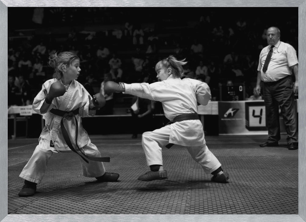 A black and white photograph of two young girls in a karate competition. They are wearing white gis, black belts, and boxing gloves. One girl is in the middle of throwing a punch at her opponent. In the background, a referee stands watching the match, and spectators are blurred in the stands. The image is presented within a silver-colored frame. Print